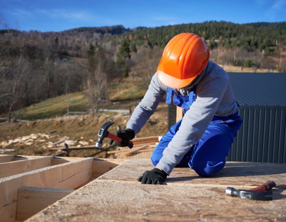 Carpenter hammering nail into OSB panel on the roof top of future cottage. Man worker building wooden frame house. Carpentry and construction concept.